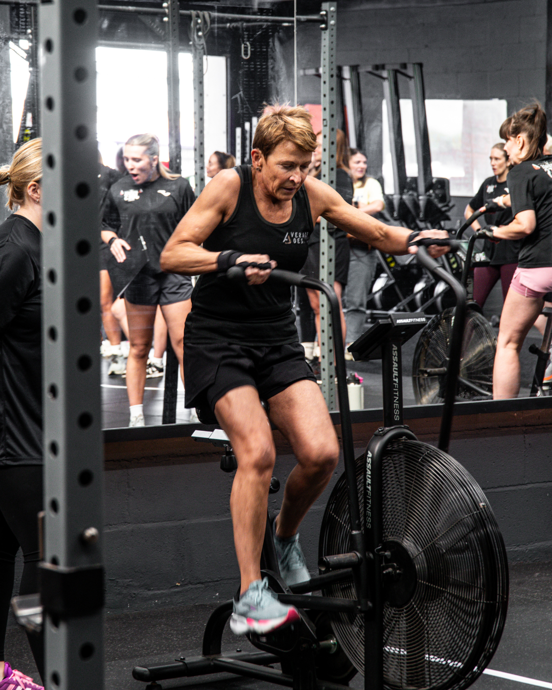 Older woman using an assault bike during a fitness competition at a modern Gateshead gym, pushing through an intense workout in a supportive group environment