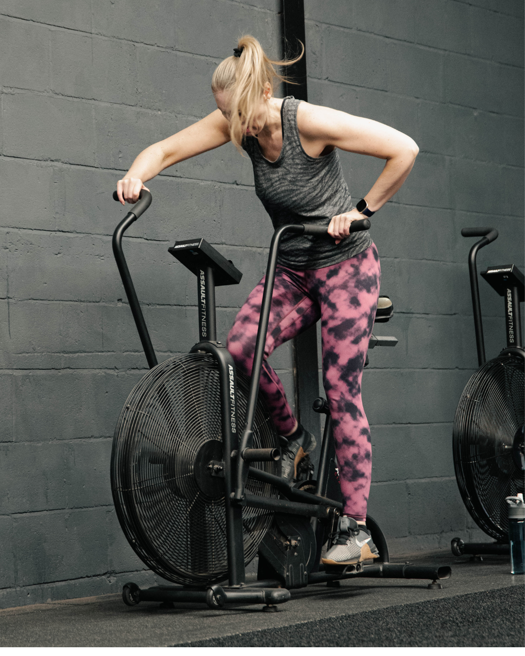 Woman using an assault bike during a group fitness class in a modern gym, pushing through a high-energy cardio workout alongside other members