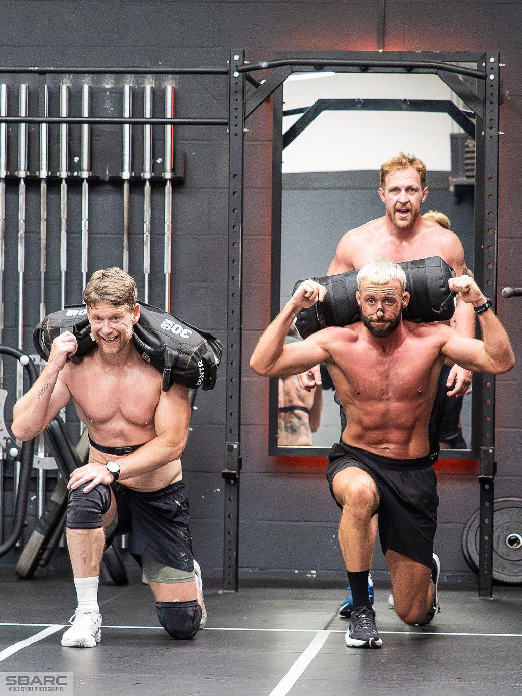 Men performing sandbag lunges during a HYROX-style training event at a Gateshead gym, pushing through a functional fitness workout in a high-energy environment